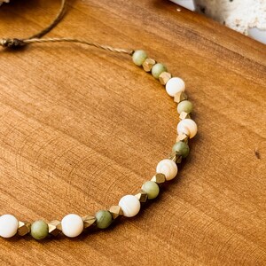 May include: A beaded bracelet featuring alternating round white beads, round sage green beads, and small metallic gold-toned faceted cube spacers. The beads are strung on a thin brown cord, which extends from the bracelet in a simple adjustable tie.