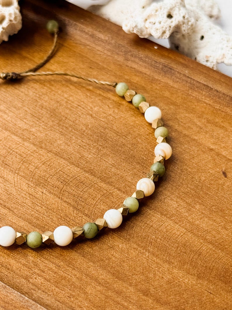 May include: A beaded bracelet featuring alternating round white beads, round sage green beads, and small metallic gold-toned faceted cube spacers. The beads are strung on a thin brown cord, which extends from the bracelet in a simple adjustable tie.