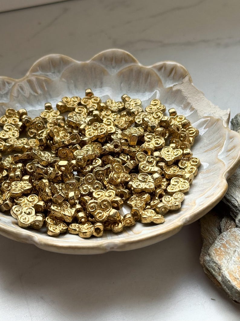 May include: A bowl filled with gold-colored metal beads. Each bead is shaped like a stylized flower with a hole in the center for stringing.