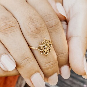 K&ouml;nnte beinhalten: Ein goldener Ring mit einem filigranen, kunstvollen Design, das ein Blumenmotiv zeigt. Der Ring wird am Finger einer Person mit heller Haut getragen.