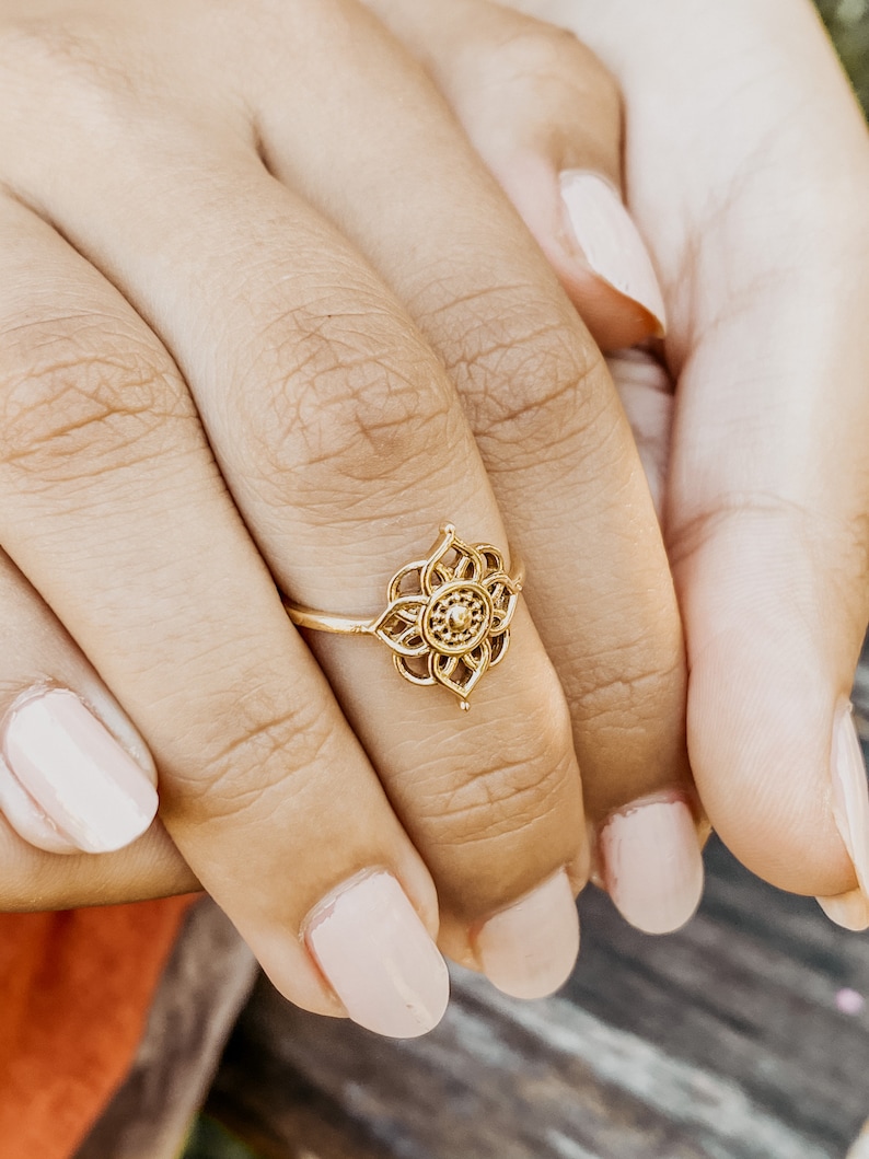 K&ouml;nnte beinhalten: Ein goldener Ring mit einem filigranen, kunstvollen Design, das ein Blumenmotiv zeigt. Der Ring wird am Finger einer Person mit heller Haut getragen.