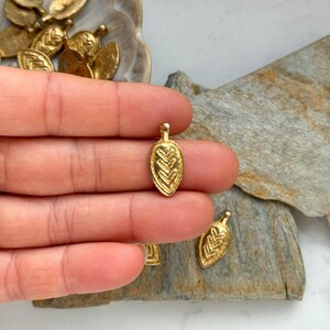 May include: A close-up of a gold leaf charm with a detailed design. The charm is small and delicate, and it would be perfect for adding a touch of elegance to jewelry or crafts.