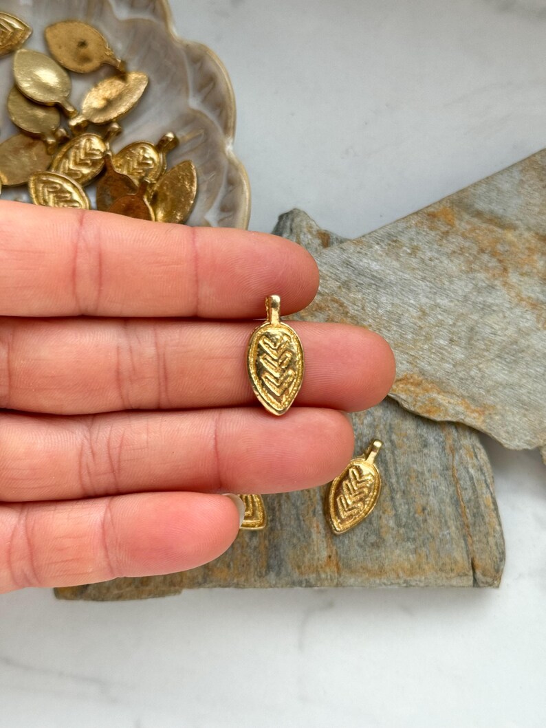 May include: A close-up of a gold leaf charm with a detailed design. The charm is small and delicate, and it would be perfect for adding a touch of elegance to jewelry or crafts.