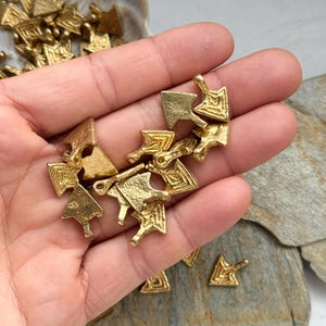 May include: A hand holding a pile of gold-colored metal arrow charms. The charms are intricately designed with geometric patterns and have a slightly textured surface.