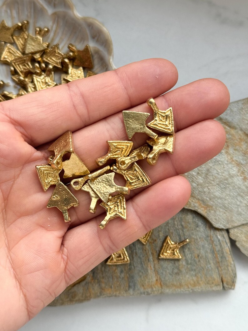 May include: A hand holding a pile of gold-colored metal arrow charms. The charms are intricately designed with geometric patterns and have a slightly textured surface.