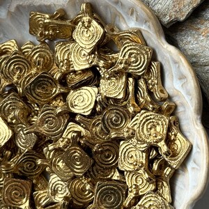 K&ouml;nnte beinhalten: Eine Schale gef&uuml;llt mit goldfarbenen Metall-Anh&auml;ngern. Jeder Anh&auml;nger ist ein Quadrat mit einem Spiralmuster.