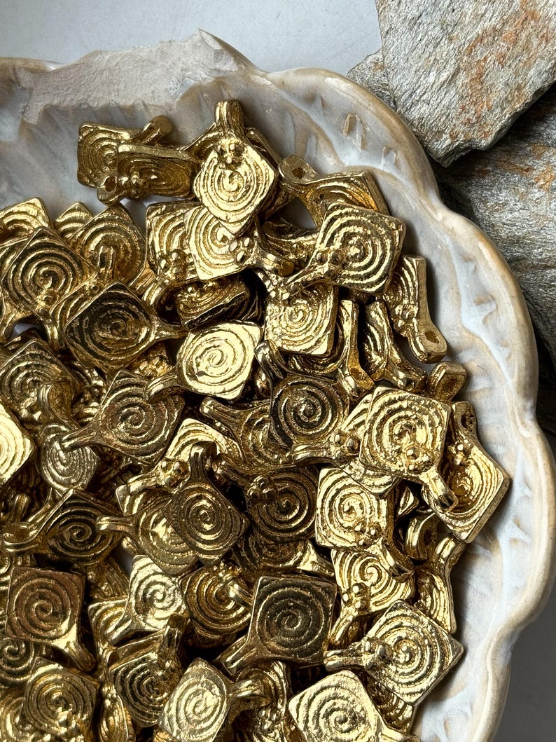 K&ouml;nnte beinhalten: Eine Schale gef&uuml;llt mit goldfarbenen Metall-Anh&auml;ngern. Jeder Anh&auml;nger ist ein Quadrat mit einem Spiralmuster.