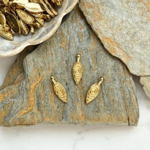 May include: Three gold leaf-shaped charms with a swirl design. The charms are on a gray stone surface.