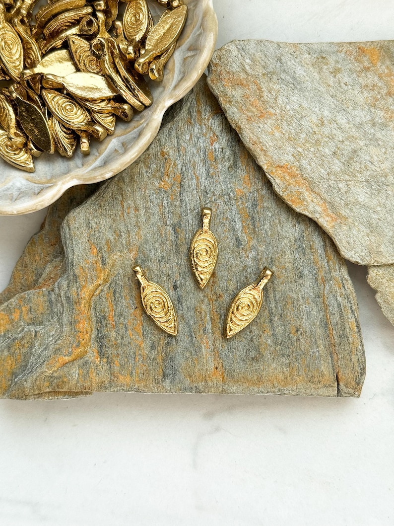 May include: Three gold leaf-shaped charms with a swirl design. The charms are on a gray stone surface.