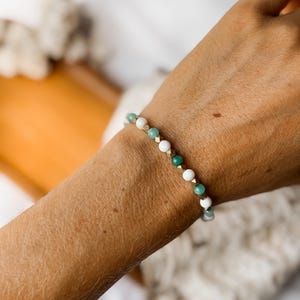 Puede incluir: Una pulsera de cuentas que presenta una alternancia de cuentas redondas blancas, cuentas redondas de color verde transl&uacute;cido y peque&ntilde;as cuentas espaciadoras met&aacute;licas de tono dorado. La pulsera sigue un patr&oacute;n repetitivo de estos tres tipos de cuentas.