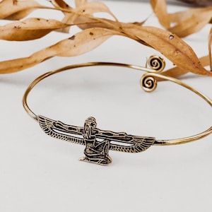 May include: A gold-toned metal cuff bracelet featuring an Egyptian winged goddess design. The bracelet has a slender band with spiral accents and a detailed central figure with outstretched wings. The background includes dried leaves.