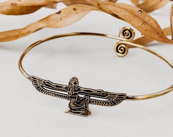 Puede incluir: Una pulsera de brazalete de metal dorado con un diseño de diosa egipcia alada. La pulsera tiene una banda delgada con detalles en espiral y una figura central detallada con alas extendidas. El fondo incluye hojas secas.