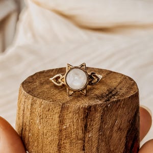 Puede incluir: Un anillo dorado con un diseño de estrella alrededor de una piedra preciosa redonda y blanca. El anillo se exhibe sobre un pequeño bloque de madera redondo. El diseño del anillo incluye pequeños adornos decorativos a ambos lados de la estrella.