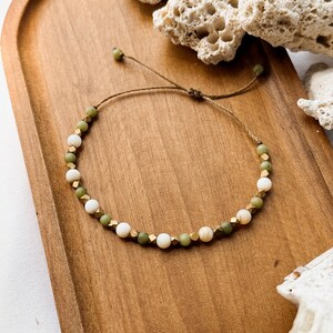 May include: A beaded bracelet featuring alternating round white beads, round olive green beads, and small metallic gold-toned faceted cube spacers. The bracelet is strung on a thin brown cord with an adjustable sliding knot closure mechanism.