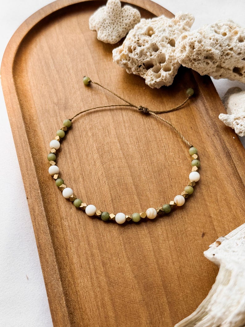 May include: A beaded bracelet featuring alternating round white beads, round olive green beads, and small metallic gold-toned faceted cube spacers. The bracelet is strung on a thin brown cord with an adjustable sliding knot closure mechanism.