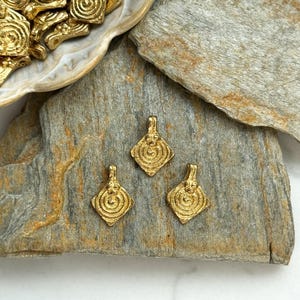 K&ouml;nnte beinhalten: Drei goldfarbene Metallanh&auml;nger mit geometrischem Design. Die Anh&auml;nger sind diamantf&ouml;rmig mit einem kreisf&ouml;rmigen Muster in der Mitte. Die Anh&auml;nger liegen auf einer grauen Steinoberfl&auml;che.