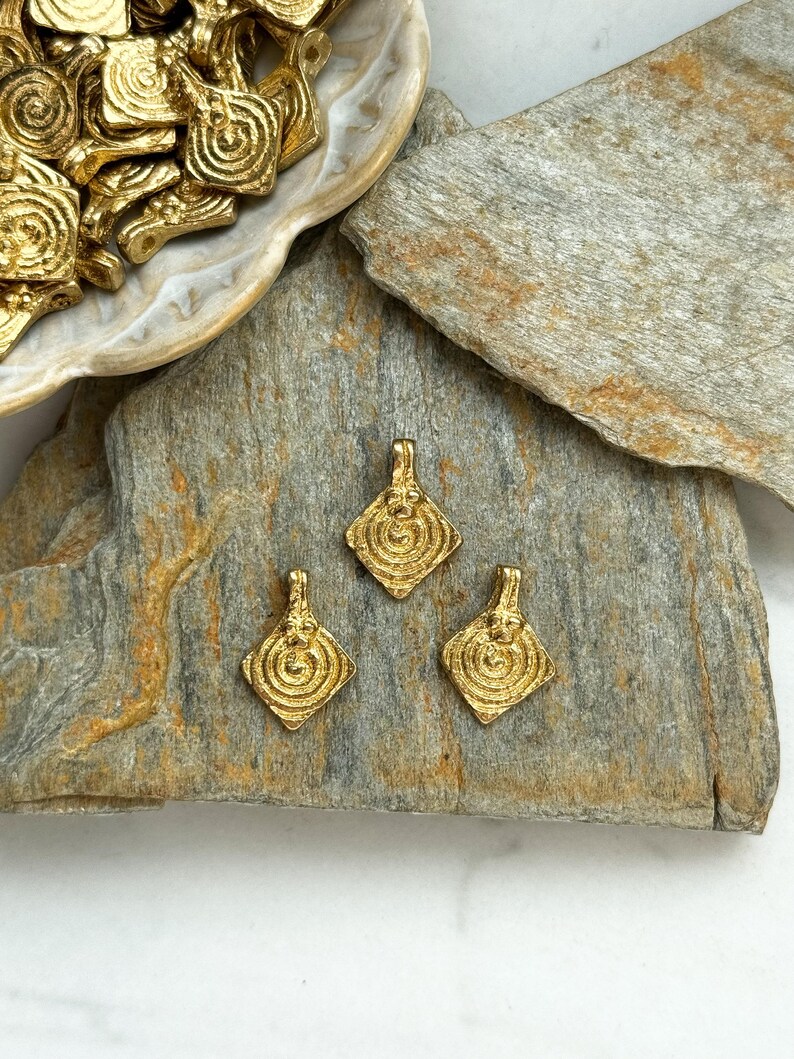 K&ouml;nnte beinhalten: Drei goldfarbene Metallanh&auml;nger mit geometrischem Design. Die Anh&auml;nger sind diamantf&ouml;rmig mit einem kreisf&ouml;rmigen Muster in der Mitte. Die Anh&auml;nger liegen auf einer grauen Steinoberfl&auml;che.
