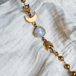 May include: A gold-toned chain necklace featuring a series of small spherical beads, two crescent moon charms, and a central round, iridescent moonstone cabochon. The metal components have a matte finish, and the design is arranged in a linear pattern.