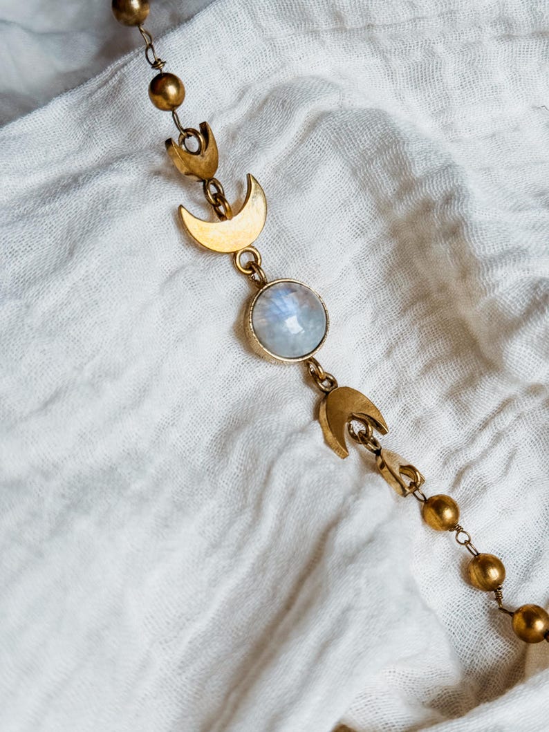 May include: A gold-toned chain necklace featuring a series of small spherical beads, two crescent moon charms, and a central round, iridescent moonstone cabochon. The metal components have a matte finish, and the design is arranged in a linear pattern.