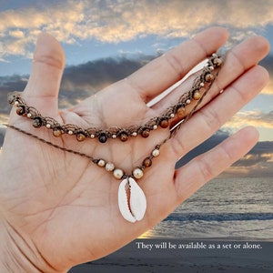 May include: A brown and beige beaded necklace with a cowrie shell pendant. The necklace is held in a hand against a background of a blue sky and ocean.