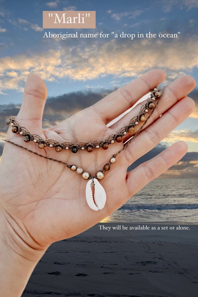 May include: A brown and beige beaded necklace with a cowrie shell pendant. The necklace is held in a hand against a background of a blue sky and ocean.