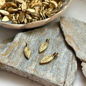 May include: Three gold leaf-shaped pendants with a swirl design. The pendants are on a gray stone surface.