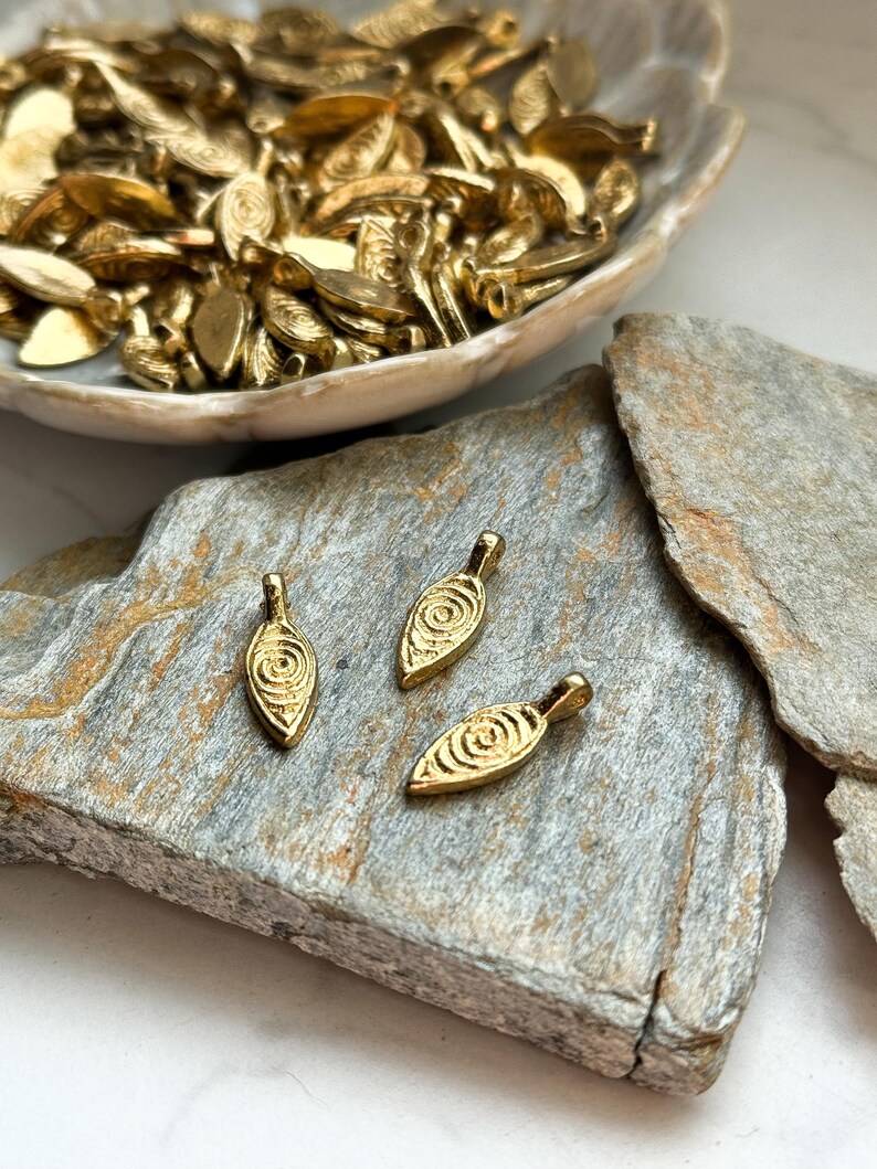 May include: Three gold leaf-shaped pendants with a swirl design. The pendants are on a gray stone surface.