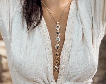 May include: A gold-colored pendant necklace with multiple decorative elements. The necklace features a series of circular and floral designs, each with a central stone. The necklace is worn over a white, textured top.