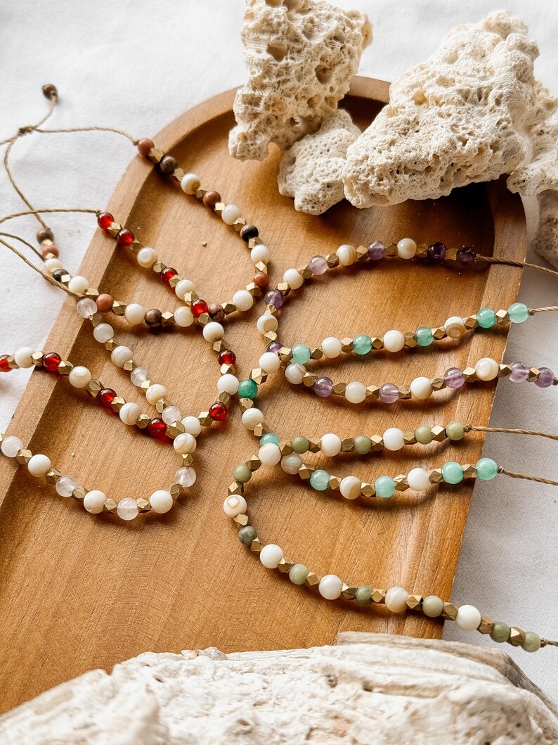 Puede incluir: Una colecci&oacute;n de pulseras ajustables hechas de cuerda natural, con un patr&oacute;n repetitivo de cuentas redondas blancas, espaciadores met&aacute;licos dorados facetados y varias cuentas de piedras preciosas de colores, incluyendo rojas, verdes, moradas y claras.
