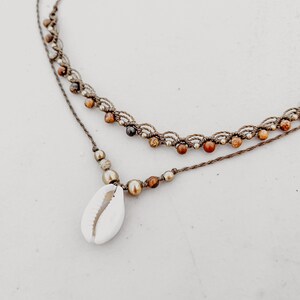 May include: A layered necklace with brown cord and small brown, gold, and red beads. The necklace has a cowrie shell pendant.