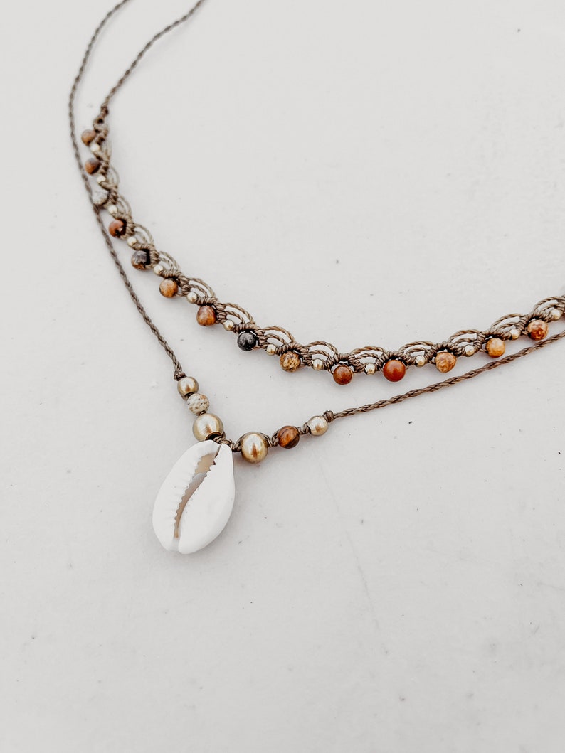 May include: A layered necklace with brown cord and small brown, gold, and red beads. The necklace has a cowrie shell pendant.
