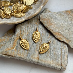 May include: Three gold leaf-shaped charms with a geometric pattern. The charms are on a piece of wood.