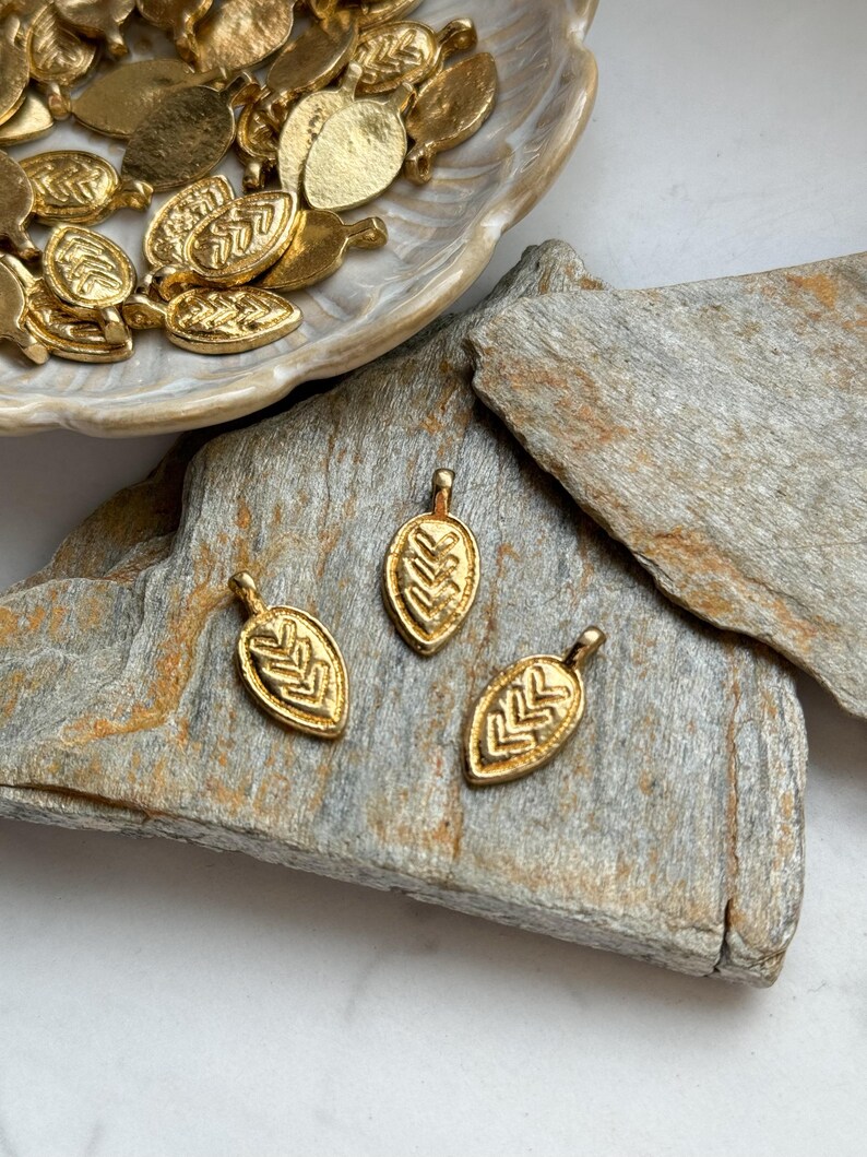 May include: Three gold leaf-shaped charms with a geometric pattern. The charms are on a piece of wood.