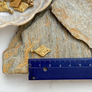 Pode incluir: Pingente dourado em forma de losango com um design ornamentado, com cerca de 2,5 cm de comprimento. V&aacute;rios pingentes semelhantes s&atilde;o vis&iacute;veis em uma tigela decorativa. O pingente est&aacute; em uma superf&iacute;cie de pedra natural.