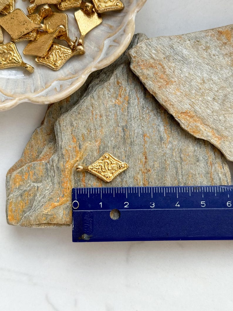Pode incluir: Pingente dourado em forma de losango com um design ornamentado, com cerca de 2,5 cm de comprimento. V&aacute;rios pingentes semelhantes s&atilde;o vis&iacute;veis em uma tigela decorativa. O pingente est&aacute; em uma superf&iacute;cie de pedra natural.