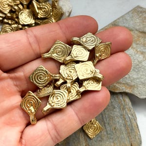 K&ouml;nnte beinhalten: Ein Haufen goldfarbener Metall-Anh&auml;nger mit einem Spiralmuster. Die Anh&auml;nger sind quadratisch mit einer kleinen Schlaufe oben.