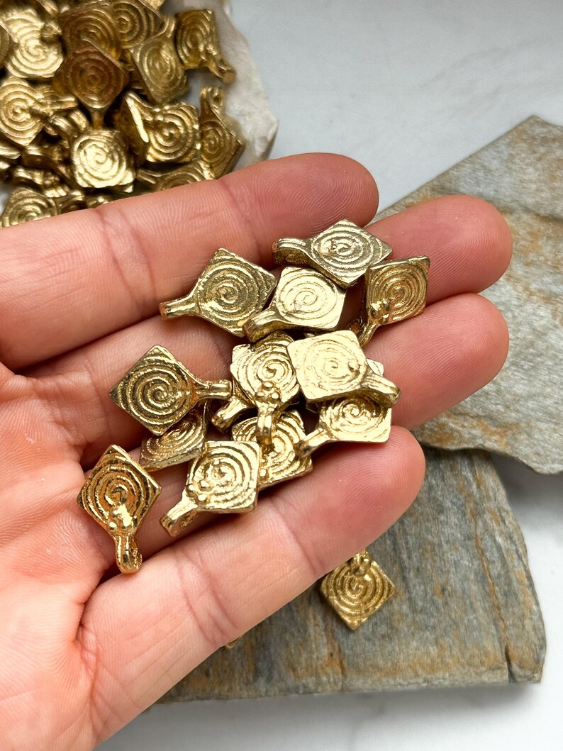 K&ouml;nnte beinhalten: Ein Haufen goldfarbener Metall-Anh&auml;nger mit einem Spiralmuster. Die Anh&auml;nger sind quadratisch mit einer kleinen Schlaufe oben.