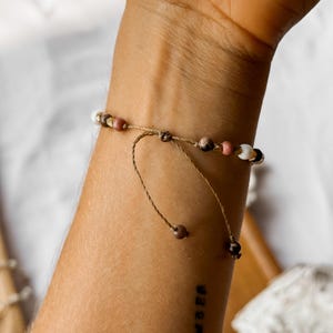 May include: A delicate adjustable bracelet featuring a thin brown cord with small beads in shades of white, pink, brown, and metallic gold. The bracelet includes two dangling cord ends, each finished with a single small brown bead for a secure fit.