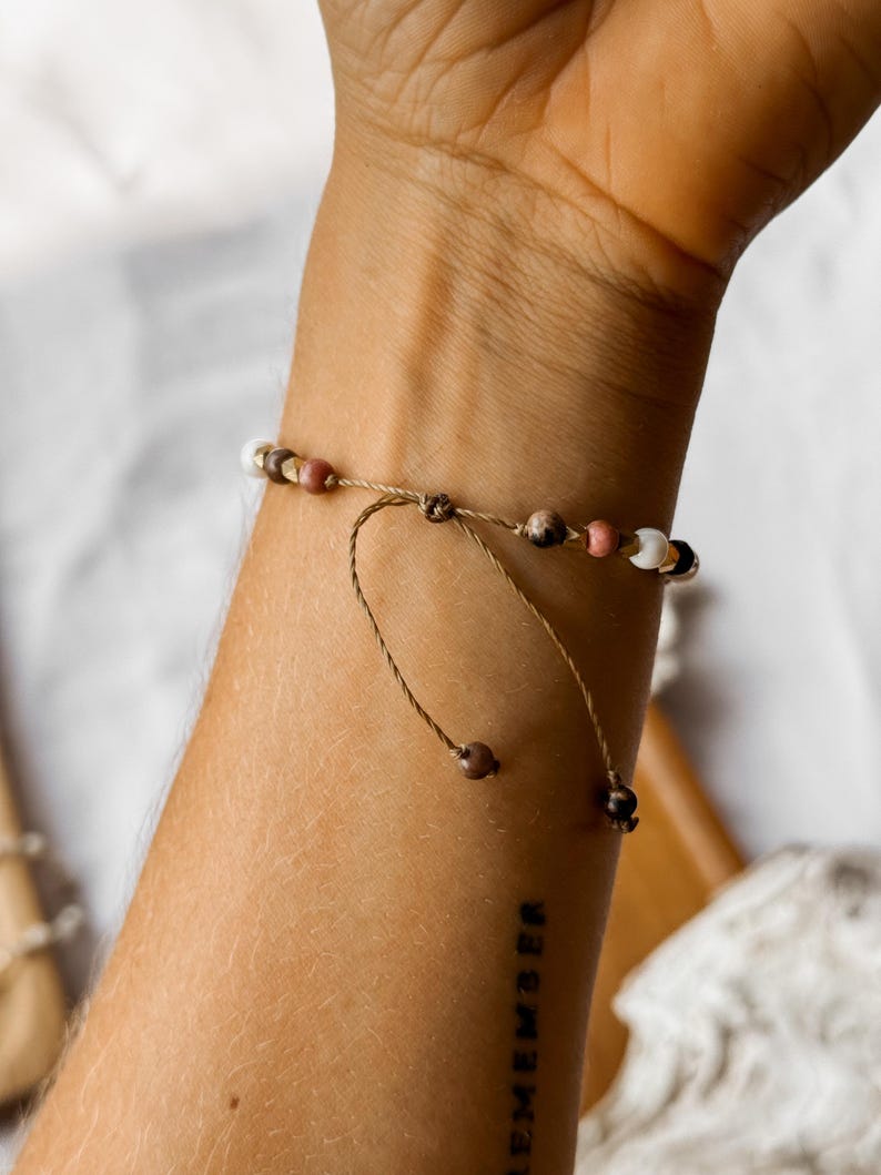 May include: A delicate adjustable bracelet featuring a thin brown cord with small beads in shades of white, pink, brown, and metallic gold. The bracelet includes two dangling cord ends, each finished with a single small brown bead for a secure fit.