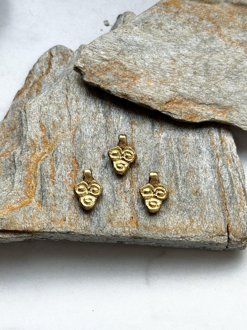 May include: Three gold-colored metal charms in the shape of a cluster of grapes. The charms are on a rough, gray, textured surface.