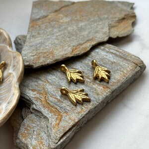 以下が含まれることがあります： 灰色と茶色の石の表面に、金色の葉の形をしたチャームが4つあります。チャームは小さく繊細で、シンプルなデザインです。