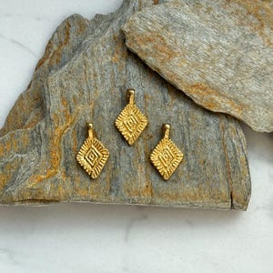 Puede incluir: Tres abalorios de metal de color dorado con un dise&ntilde;o geom&eacute;trico. Los abalorios est&aacute;n dispuestos sobre una superficie de piedra gris y marr&oacute;n.