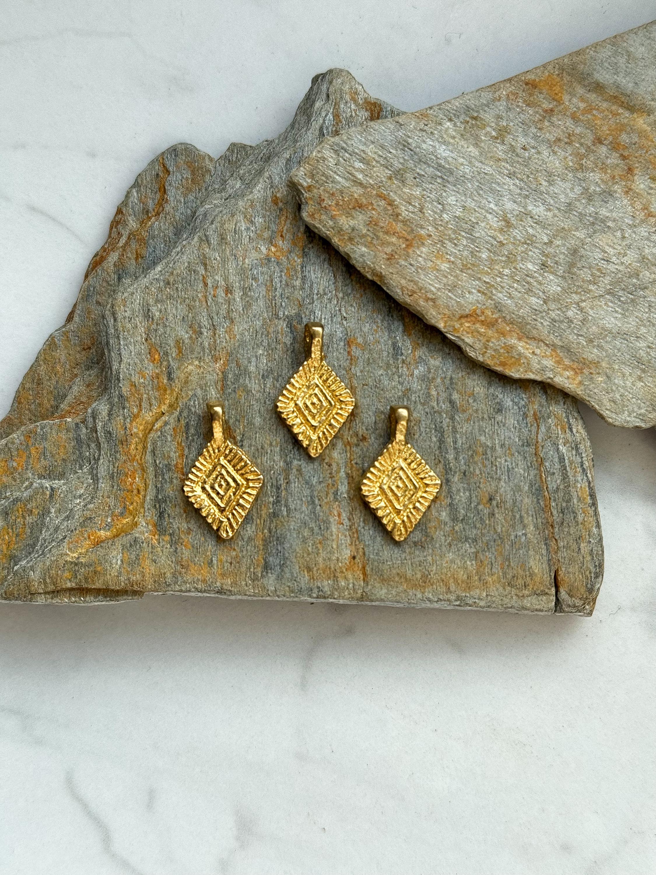 Puede incluir: Tres abalorios de metal de color dorado con un dise&ntilde;o geom&eacute;trico. Los abalorios est&aacute;n dispuestos sobre una superficie de piedra gris y marr&oacute;n.