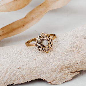 Puede incluir: Un anillo de oro con un delicado diseño de filigrana y una piedra blanca en el centro. El anillo está sobre una superficie de madera marrón claro.
