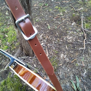 May include: A brown leather guitar strap with a silver buckle. The strap is attached to a wooden guitar.