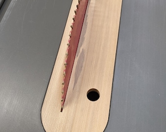 Puede incluir: Una placa de inserción para sierra de mesa de madera clara con forma rectangular redondeada, que presenta una ranura central para una hoja de sierra circular roja y una cuña separadora. La placa incluye un pequeño orificio circular y el texto Pixels Production.
