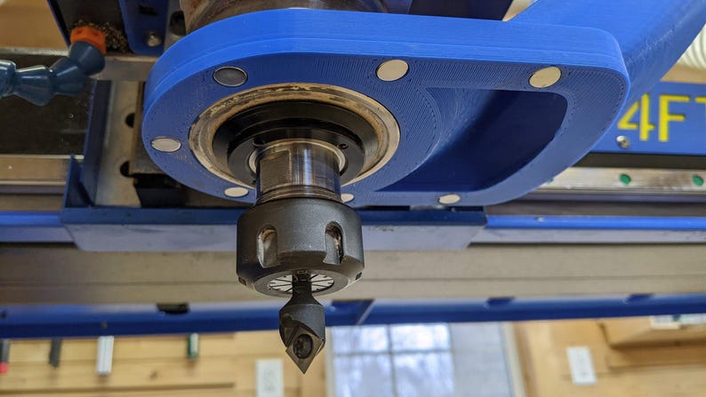 May include: Close-up of a blue and black CNC router with a metal cutting bit attached to the spindle. The router is mounted on a metal frame and has a blue plastic cover.