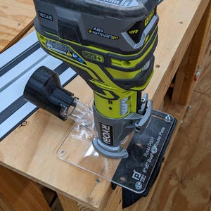 Puede incluir: Una fresadora inalámbrica Ryobi de 18V, negra y amarilla, montada en un banco de trabajo de madera. Tiene una base de plástico transparente y un accesorio de recogida de polvo negro. El texto "Ryobi P601 6"x6" Bush Router Plate" es visible.