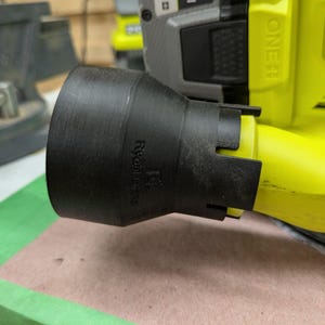May include: A black plastic dust collection adapter attached to a lime green power sander. The adapter features a flared circular opening and the embossed text Ryobi Corner. The sander body displays the text PCL406 18V 5 in. (127 mm).