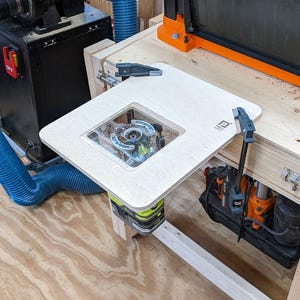 Puede incluir: Una mesa de enrutador de madera blanca con un inserto de plástico transparente para un enrutador. La mesa está unida a una superficie de madera más grande con abrazaderas. La mesa de enrutador está sobre un suelo de madera.